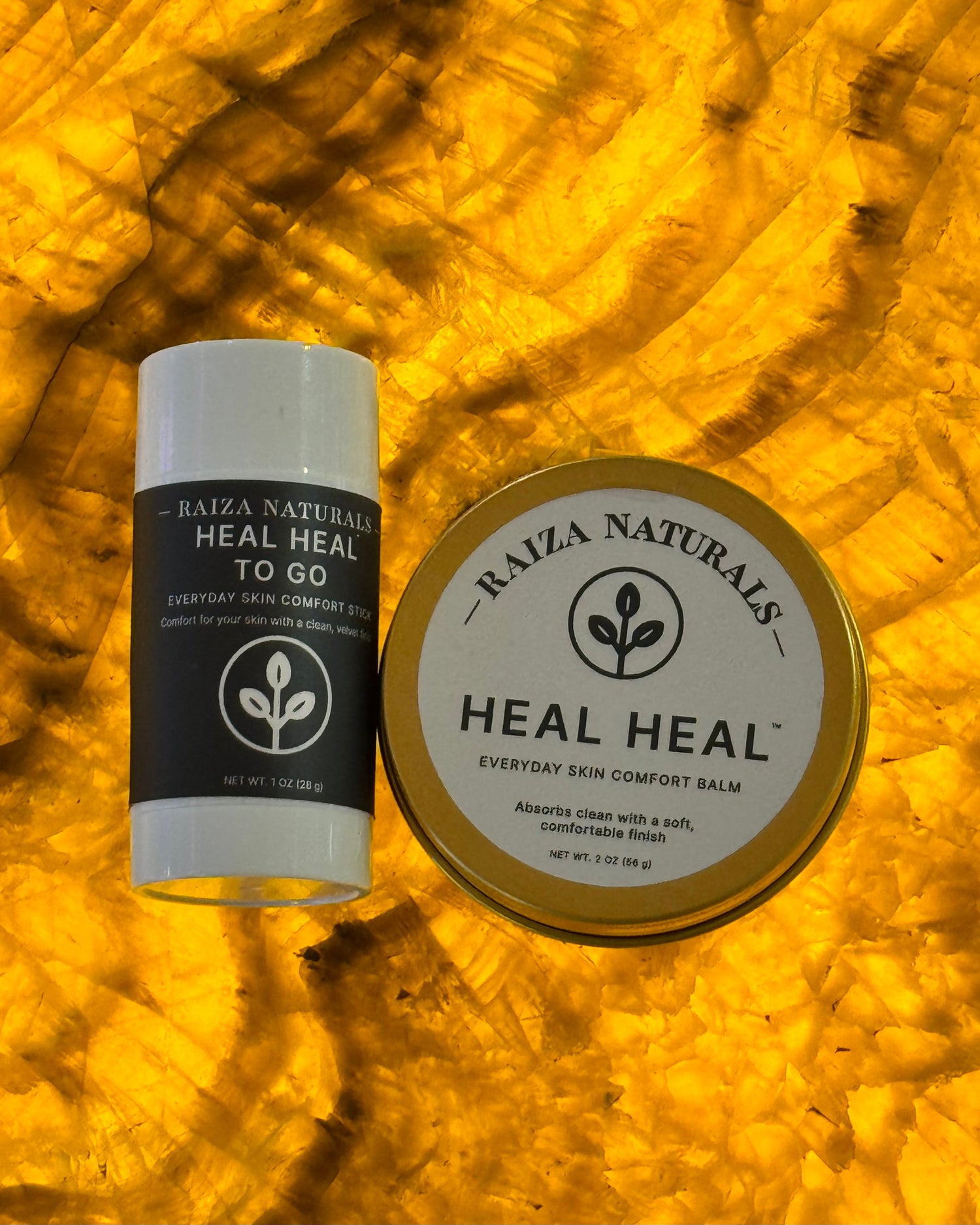 Raiza Naturals Heal Heal balm and Heal Heal To Go skin comfort stick displayed on a warm golden stone background.