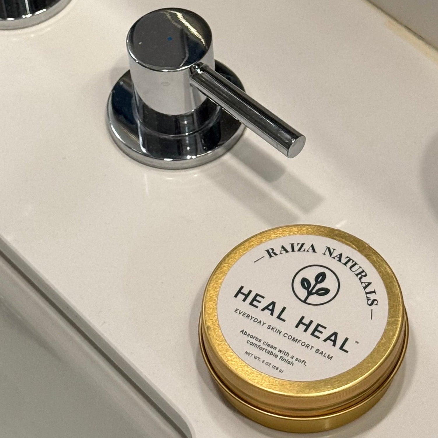 Heal Heal Everyday Skin Comfort Balm in a 2 oz gold tin beside a bathroom sink, ready for everyday use.